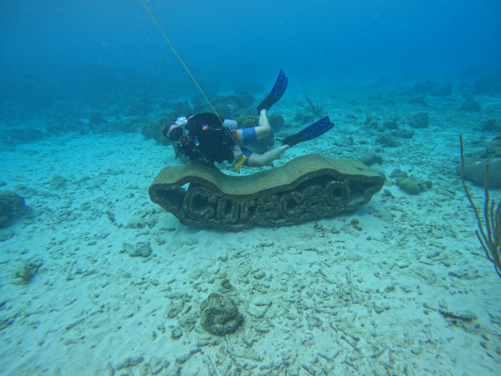Scuba diver posing underwater next to a large stone sculpture that spells out "Curaçao" on the sandy ocean floor at the LionsDive House Reef.